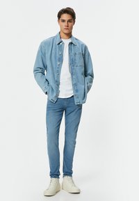 Chaqueta de denim azul claro con un bolsillo frontal, superpuesta a una camiseta blanca. Combinada con jeans ajustados azul claro y zapatillas blancas de caña alta.