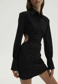 Svart skjortklänning med långa ärmar, med sidoutskärningar, figurnära midja och knä-lång fåll. Tillverkad av ett slätt tyg.