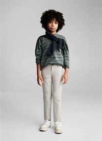 Kind in een gestreepte groen-grijze lange mouw shirt, beige broek en witte sneakers met een donkere gebreide sjaal om de nek.