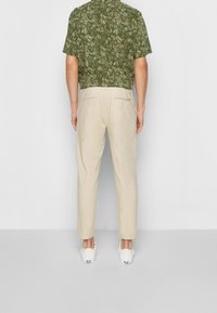 Camicia verde a maniche corte con un motivo a foglia abbinata a pantaloni beige chiaro a figura affusolata e sneakers bianche, vista da dietro.