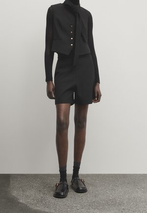 Mannequin portant un top noir à manches longues, un gilet sans manches ajusté avec des boutons dorés, un short taille haute, des chaussettes noires et des mocassins noirs.