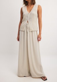 Gilet sans manches beige tricoté avec un décolleté en V profond, associé à un pantalon léger gris clair à taille élastique.