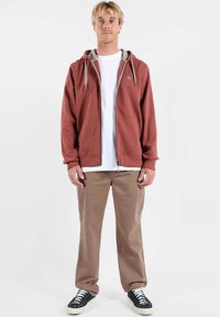 Sweat à capuche rouge à fermeture éclair avec une capuche doublée de gris, porté sur un t-shirt blanc, associé à un pantalon beige et des baskets noires. Design simple avec des poches avant.