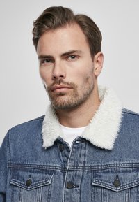 Giacca di jeans di un blu medio con colletto in finta shearling bianco, dotata di chiusura a bottoni, due tasche sul petto e una finitura leggermente vissuta.