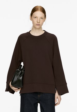 Brauner Pullover mit Raglanärmeln, geripptem Ausschnitt und lässigem Schnitt. Kombiniert mit einer kleinen schwarzen Tasche mit strukturierter Oberfläche.