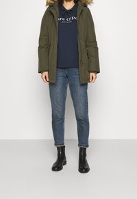 Parka verde oliva con cappuccio in pelliccia sintetica, chiusura con zip e tasche laterali; felpa navy sotto; jeans blu; stivali neri alla caviglia.