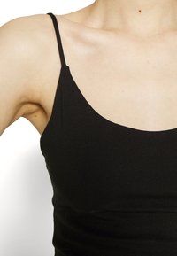 Femme portant un top noir près du corps à bretelles fines, dévoilant les épaules, le cou et une partie du haut de la poitrine sur fond blanc.