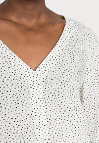 Nahaufnahme einer Brust, die eine weiße Bluse mit schwarzen unregelmäßigen Punkten und einem V-Ausschnitt trägt.