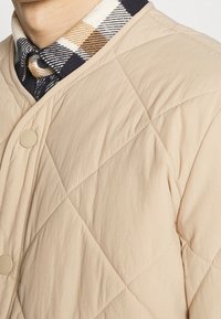 Beige gesteppte Jacke mit rundem Kragen. Der Innenkragen hat ein kariertes Muster in Marineblau, Beige und Braun.