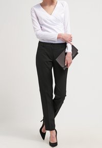 Witte lange mouw wikkeltop gecombineerd met een op maat gemaakte zwarte broek en zwarte hakken. Een rechthoekige donkere clutch maakt de outfit compleet.