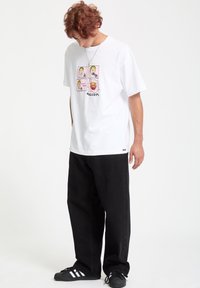 Hvid bomulds t-shirt med et farverigt grafisk print med fire tegneserieansigtter og ordet "VOLCOM." Sat sammen med sorte lige bukser.