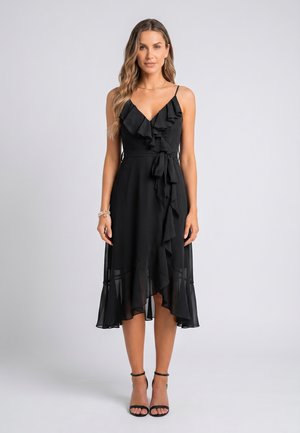 Frau trägt ein ärmelloses schwarzes Midikleid mit Rüschen und schwarze Pumps mit Knöchelriemen, steht vor einem schlichten hellgrauen Hintergrund.