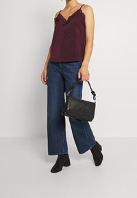 Camisola bordeaux con pizzo, abbinata a jeans a gamba larga blu e stivaletti neri con tacco block a punta aperta, mentre tiene una borsa nera dalla texture interessante.