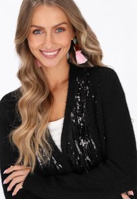 Fekete kardigán flitteres díszítéssel, redőzött kialakításban, fehér felső fölött viselve. Hosszú, hullámos haj és rózsaszín bojtos fülbevalók láthatók.