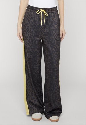 Kobieta stojąca, ubrana w szerokie spodnie w panterkę z żółtymi paskami po bokach i żółtym sznurkiem, w połączeniu z białymi trampkami.