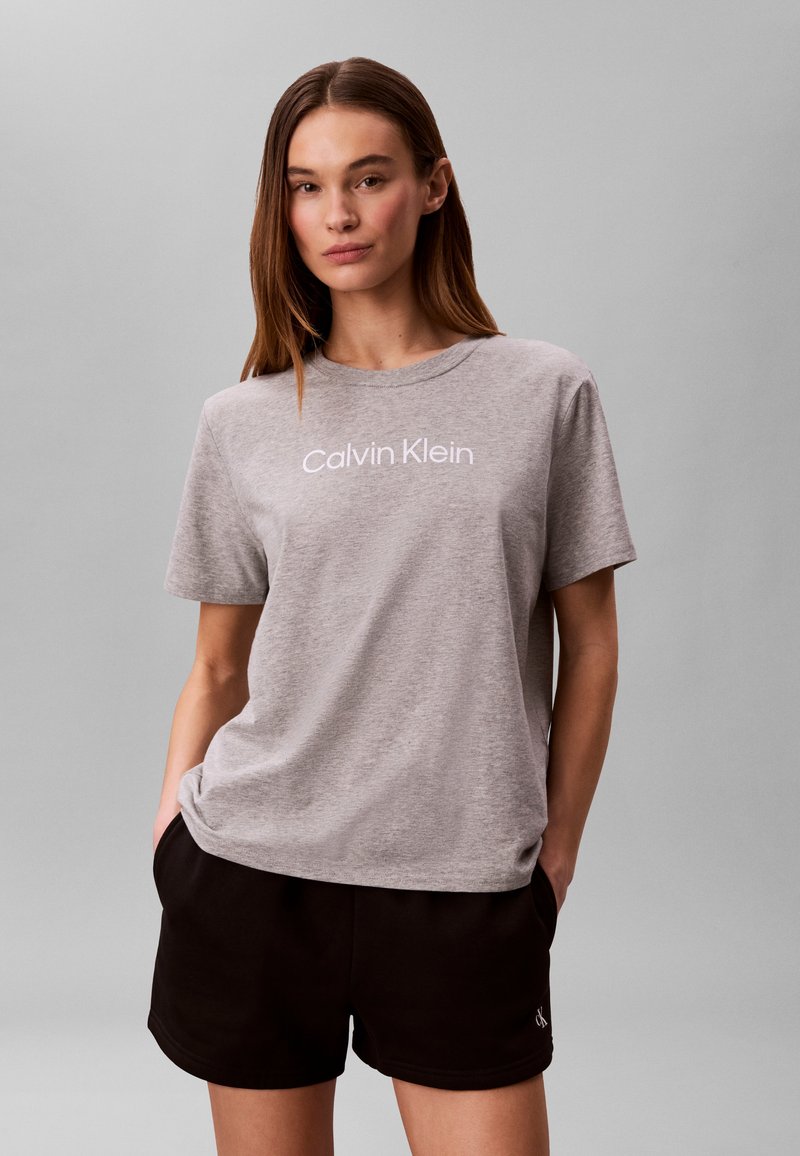 Femme portant un t-shirt gris Calvin Klein et un short noir, les mains dans les poches, sur un fond gris uni.