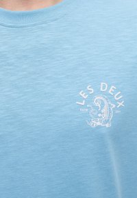 Ανοιχτό μπλε ύφασμα με λευκό κείμενο "LES DEUX" πάνω από ένα στυλιζαρισμένο σχέδιο σαύρας και "ESTD 2011" από κάτω στην περιοχή του στήθους.