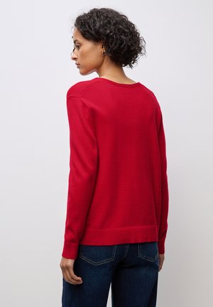 Maglione rosso in morbido tessuto, con maniche lunghe, scollatura rotonda e orlo e polsini a costine; indossato sopra jeans blu scuro.
