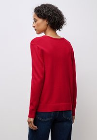 Maglione rosso in morbido tessuto, con maniche lunghe, scollatura rotonda e orlo e polsini a costine; indossato sopra jeans blu scuro.
