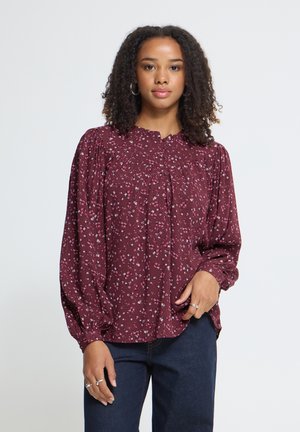 Joven mujer con cabello rizado que lleva una blusa de manga larga color granate con estampado floral pequeño y vaqueros azul oscuro sobre un fondo liso.