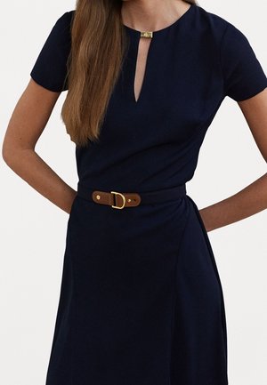 Femme portant une robe bleu marine à manches courtes avec une encolure en trou de serrure et une ceinture marron avec une boucle dorée, debout les mains sur les hanches.