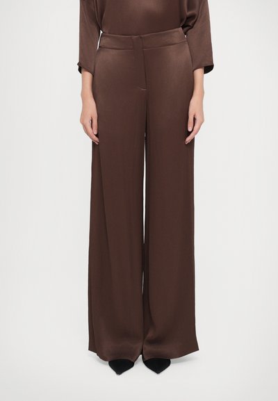 Pantalons marron à jambes larges en tissu lisse et texturé. Présente une taille haute et des plis avant subtils. Portés avec des chaussures noires à bout pointu.