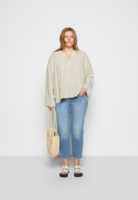 Blusa leve com riscas e decote em V, combinada com calças de ganga azuis. Modelo segura uma mala de palha e usa sandálias brancas.