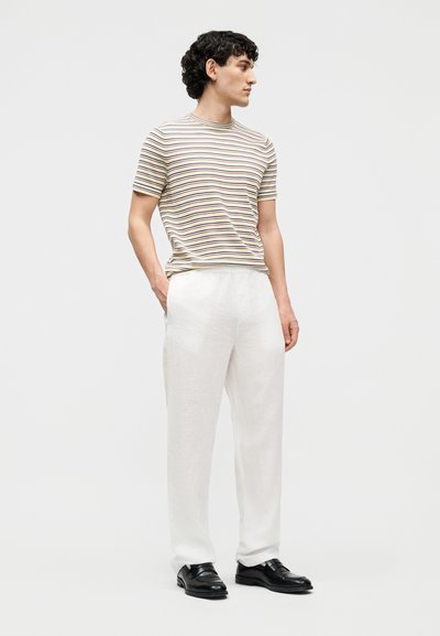 Άνδρας που φοράει κοντομάνικη μπλούζα με μπεζ και μαύρες ριγέ, άσπρο παντελόνι, μαύρα λοφτ παπούτσια, στέκεται με τα χέρια στις τσέπες, κοιτάζοντας δεξιά.