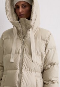 Nainen, joka pitää päällään beigeä puhallettua takkia, jossa on suuri huppu ja nyöri, vetoketju kiinni, sekä beigeä neulepipoa valkoista taustaa vasten.