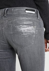 Jeans grises con un acabado brillante, que cuentan con un bolsillo trasero y una pequeña etiqueta rectangular de la marca. Textura suave con detalles de costura.