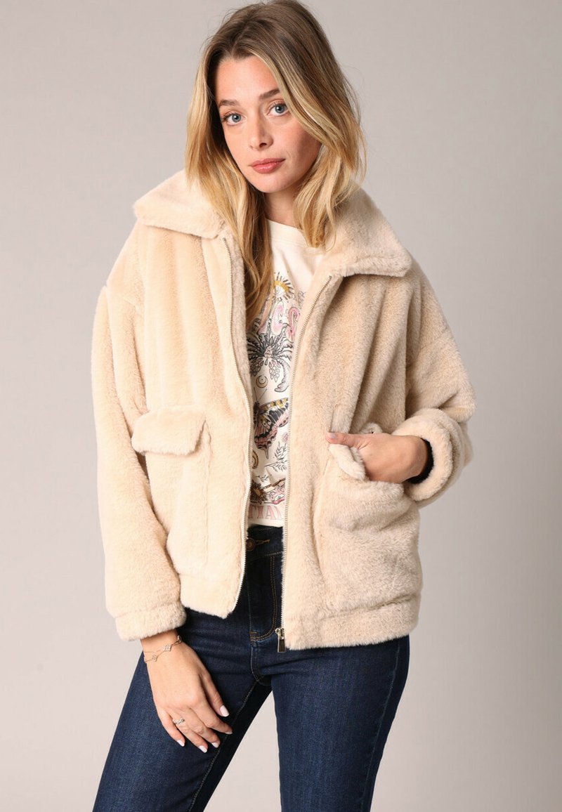 Femme blonde vêtue d'une veste zippée en fausse fourrure beige par-dessus un t-shirt graphique et un jean bleu foncé, se tenant contre un fond neutre.