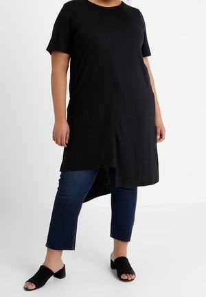 Osoba ubrana w czarną tunikę z krótkim rękawem o asymetrycznym obszyciu, nałożoną na ciemnoniebieskie, skrócone dżinsy oraz czarne klapki na słupku z odkrytymi palcami.