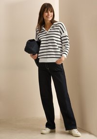 Gestreifter Strickpulli in Schwarz und Weiß, mit V-Ausschnitt; kombiniert mit dunklen Jeans und weißen Sneakers; hält eine schwarze Tasche.