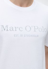 Lähikuva valkoisesta T-paidasta, jossa on haalistunut teksti "Marc O'Polo" ja "EST. IN STOCKHOLM" painettuna rintaan.