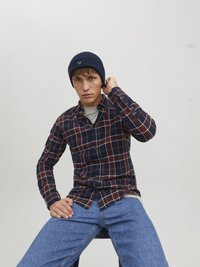 Camicia a quadri navale e rossa con bottoni, realizzata in cotone, indossata sopra una maglietta grigia. Jeans di denim blu e un berretto lavorato a maglia navy con un dettaglio del logo.