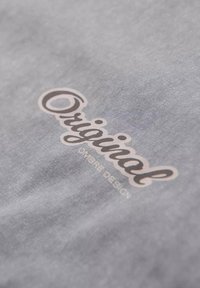 Šedá bavlněná tkanina s stylizovaným logem "Original" v světle hnědé barvě, pod nímž je menším písmem vytištěno "OMORE DESIGN". Hladká textura.