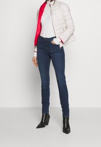 Světlá béžová prošívaná bunda s červenou podšívkou, kombinovaná s bílým žebrovaným rolákem a tmavě modrými úzkými džínami. Černé kotníkové boty na podpatku.