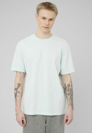 Jeune homme aux cheveux clairs portant un t-shirt bleu clair uni et un pantalon gris, montrant des tatouages floraux et de papillons sur les deux avant-bras.