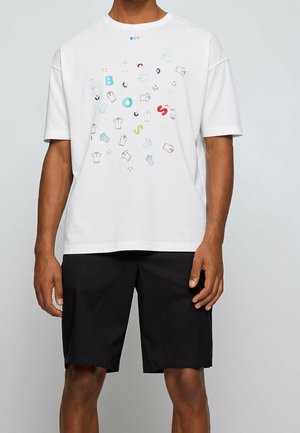 T-shirt en coton blanc avec des imprimés graphiques multicolores représentant des vêtements et des icônes. Coupe ample, col rond, associé à un short noir.