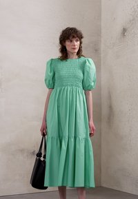 Vestido verde menta, até ao joelho, com mangas bufantes, corpete franzido e saia em camadas. Combinado com uma bolsa preta. Textura do tecido lisa.