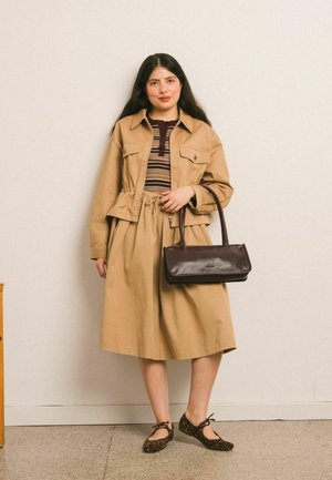 Femme debout portant une veste et une jupe couleur sable, un haut rayé, des ballerines imprimé léopard, tenant un sac à main marron foncé contre un mur uni.