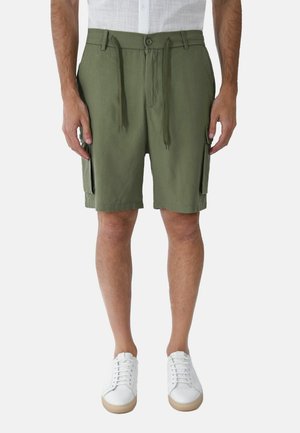 Uomo che indossa pantaloncini cargo verde oliva con bottone frontale e coulisse, abbinati a scarpe da ginnastica bianche, in piedi su uno sfondo semplice.