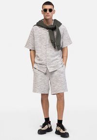 Lys grå tekstureret kortærmet skjorte og matchende shorts, suppleret af en mørkegrøn sweater draperet om halsen og sorte sneakers.