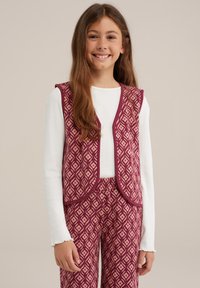 Bordeaux gekleurde patroonvest met diamantmotieven, omzoomd met effen bordeaux, gedragen over een witte blouse met lange mouwen. Bijpassende geblokte broek is zichtbaar.