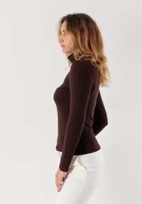 Maglia marrone a maniche lunghe con colletto alto, dal design aderente, realizzata in un tessuto liscio. Indossata con pantaloni bianchi, esposta su uno sfondo chiaro.