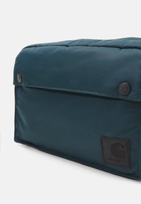 Borsa imbottita di un verde scuro con chiusura a pattina, caratterizzata da una toppa nera con logo e dettagli con bottone a scatto. Texture liscia e forma rettangolare.
