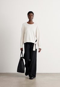 Crème sweatshirt met zijstrikdetail, gecombineerd met zwarte wijde broek en een zwarte tote bag. Glad textuur, relaxte pasvorm, minimalistisch ontwerp.