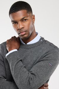 Pull en tricot gris avec une finition texturée, boutons aux épaules détaillés et un col rond classique, porté par-dessus une chemise à col bleu clair.