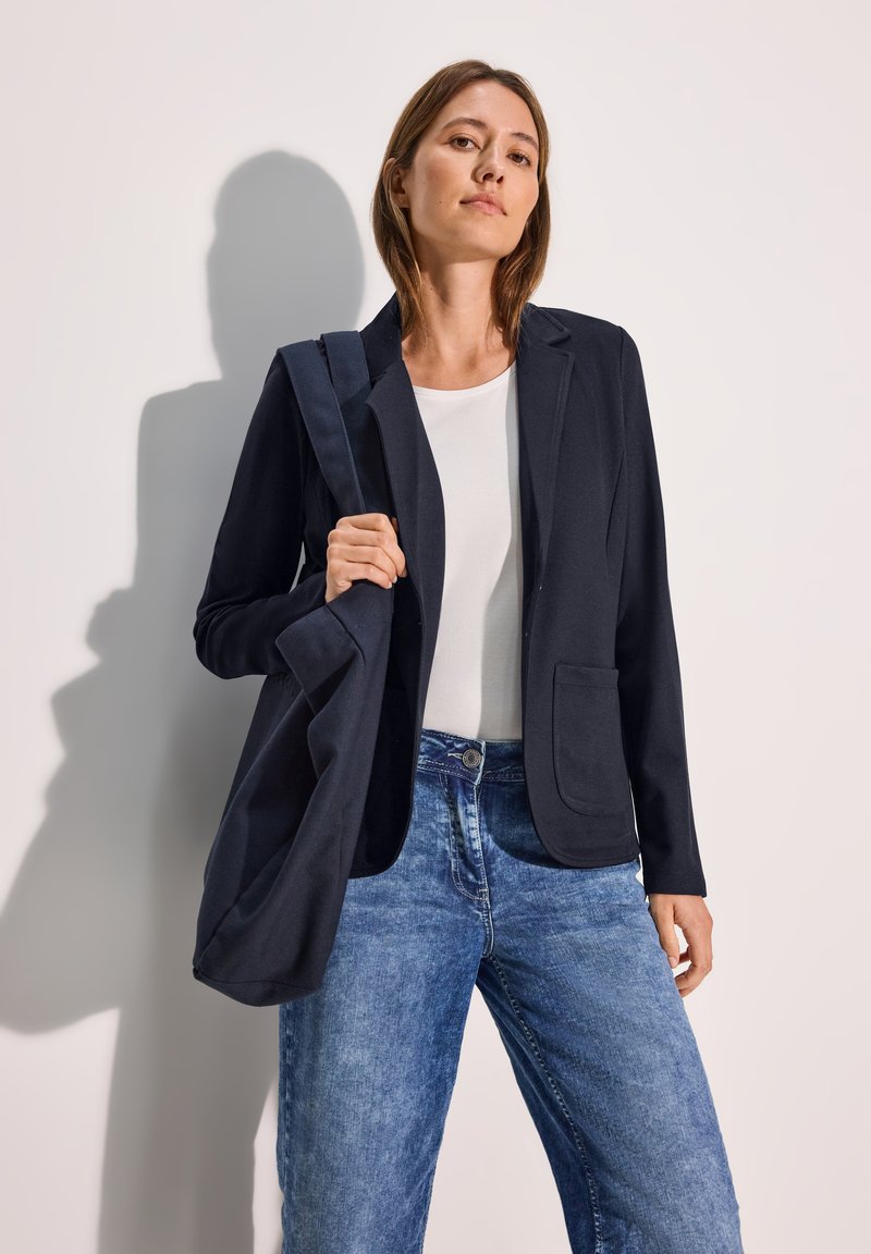 Mörkblå blazer med skräddarsydd passform och fickor, ihopparad med en vit skjorta och ljusblå vida jeans. En matchande tote-väska hänger över hennes axel.