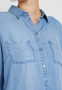 Light blue denim shirt with a collar, two chest pockets, and a button-down closure. Fabric has a soft texture and a slightly worn look.